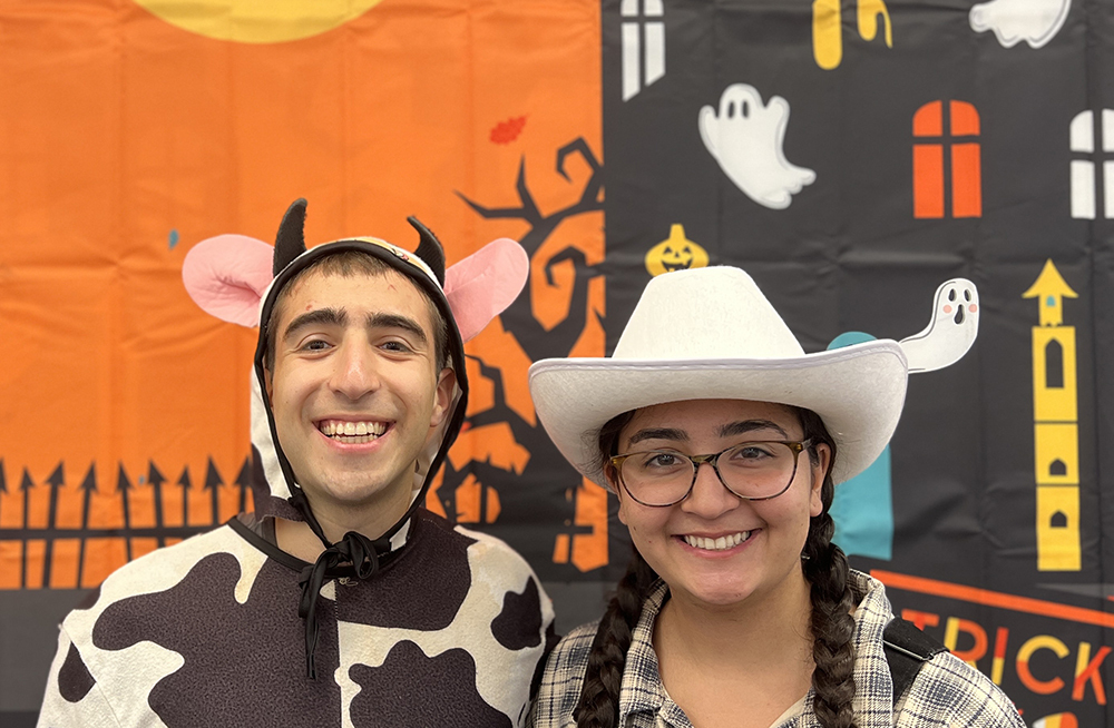 A man dressed as a cow smiles next to a woman dressed as a cowboy.