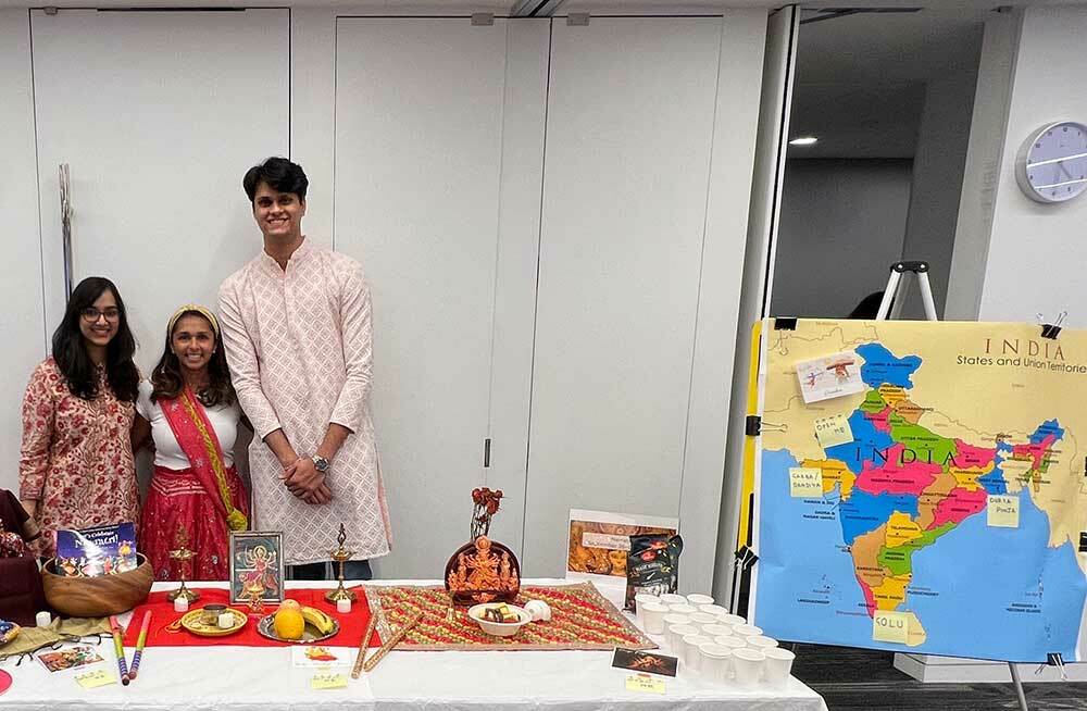 Two women and a man in Indian clothing stand behind a table with Indian food and a map of India