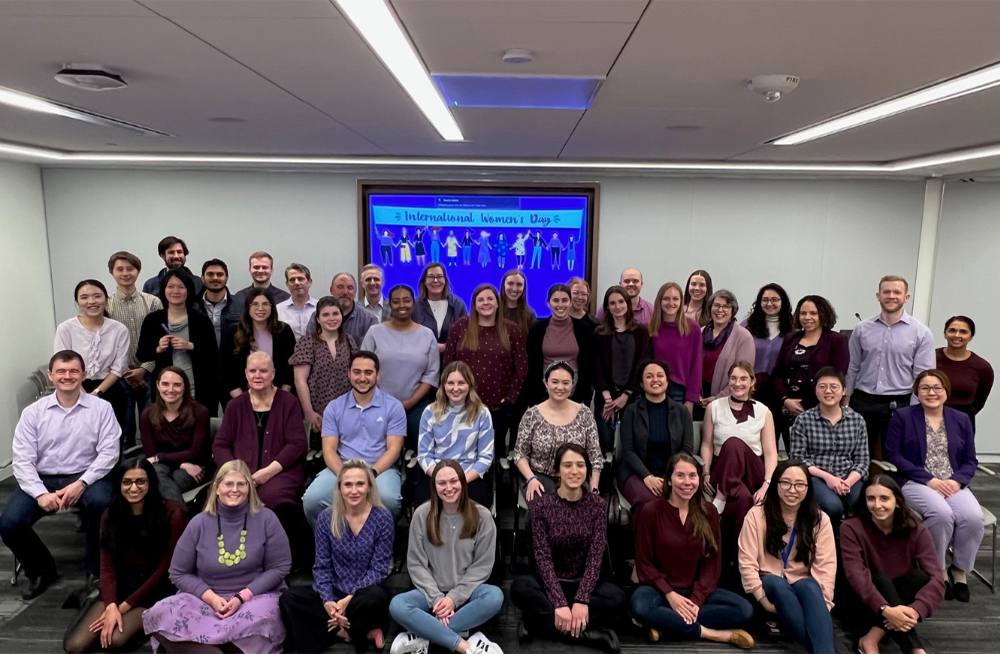 Large group of people sits in an office setting smiling at the camera and wearing different shades of purple clothing