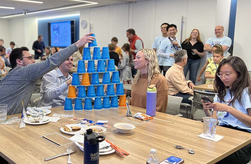 People gathered around a table stacking blue and orange plastic cups while people in the background watch and cheer