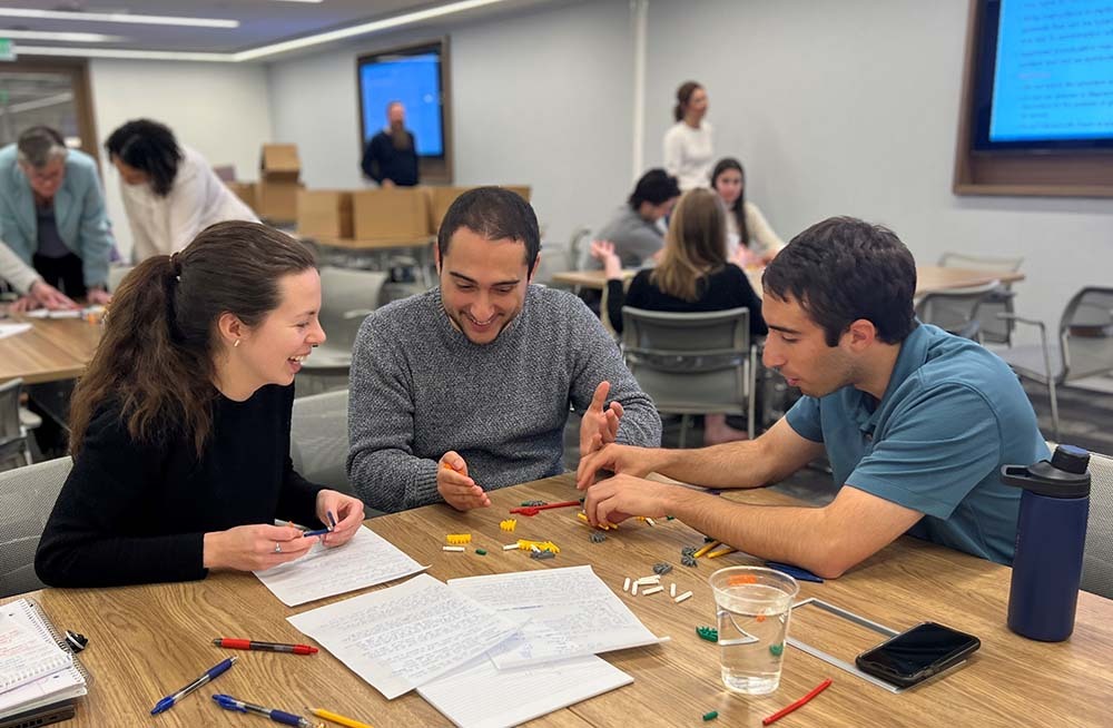 A woman with a ponytail and two men look at papers spread on a table and hold small pieces that they are trying to put together into a shape