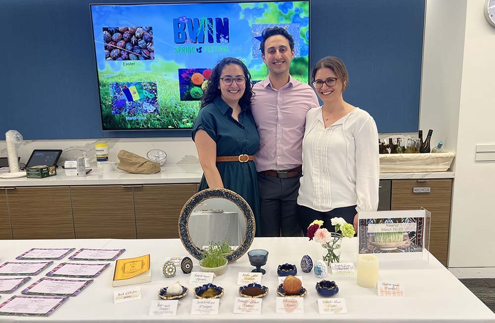 A tall man stands between two smiling women in glasses in front of a table set with different types of food in fancy dished, in front of a sign with BWIN Spring Festival