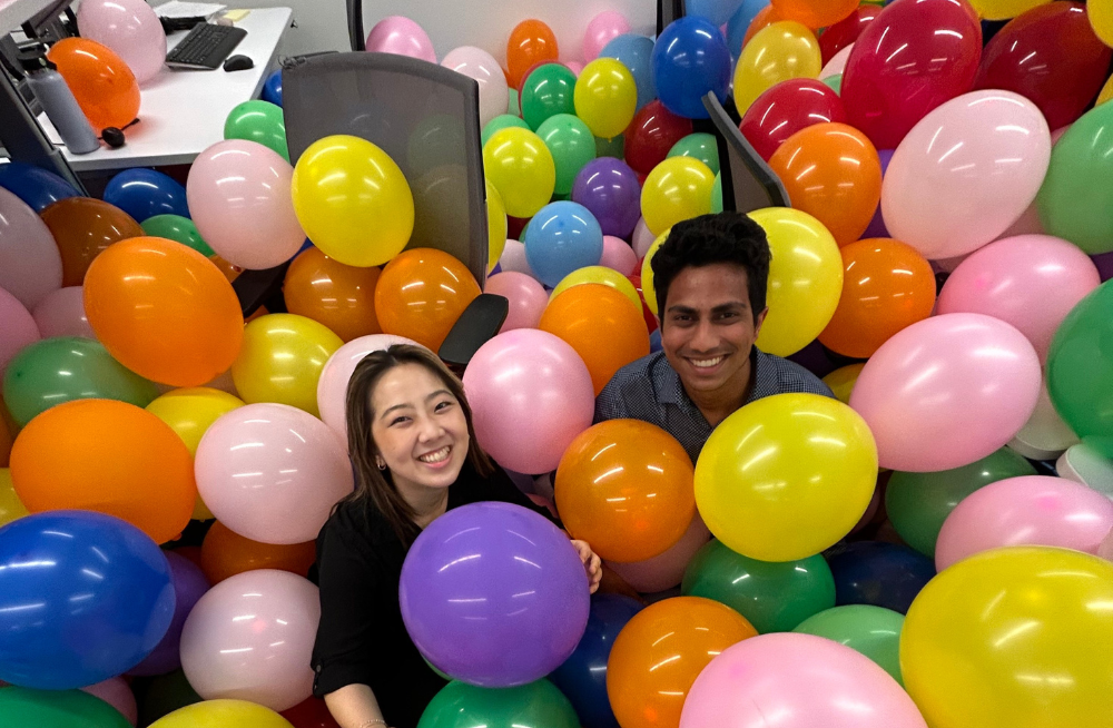One man and one women inside an office, standing waste high in balloons that fill the room; they are each holding a few balloons and smiling.
