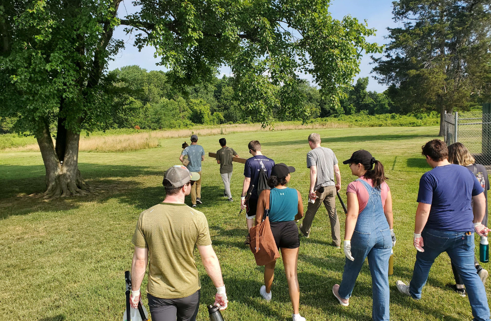 A group of adults walking together outside in casual clothing; they're walking across green grass; a clear blue sky is above them and there are trees ahead and around them.