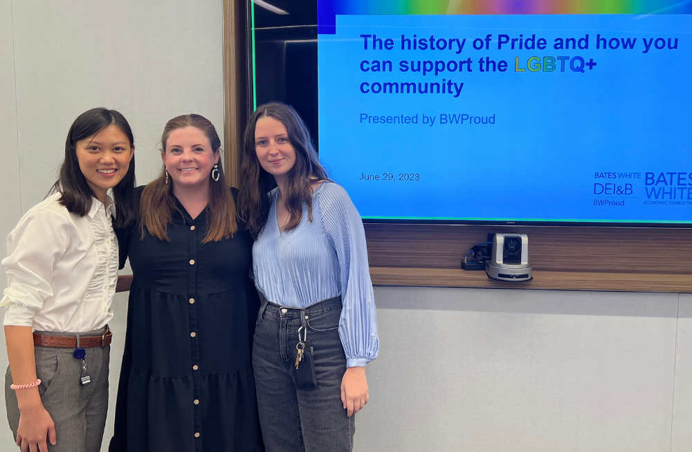 Three smiling women standing arm-and-arm together, in front of a screen featuring the title "The history of Pride and how you can support the LGBTQ+ community."