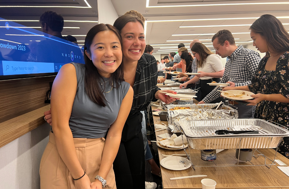 Two smiling young women stand together on one side of a buffet table. On the other side, several men and women are lined up serving themselves foods.
