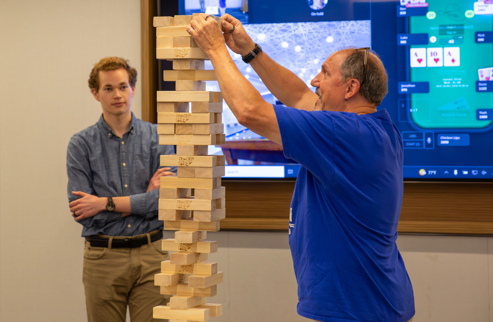A man carefully removes a block from a Jenga tower, while another man watches.