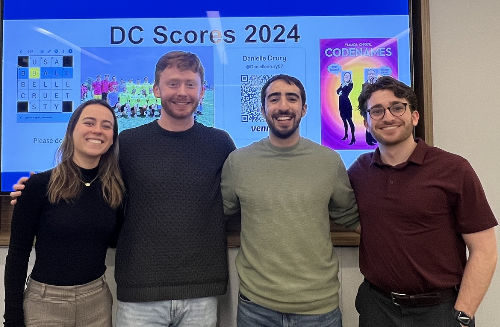 Three young men and one young woman standing together in front of a screen titled: "DC Scores 2024."