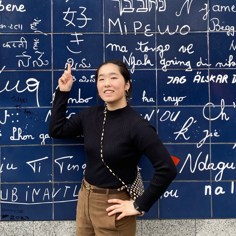 A young woman smiling and pointing at a tiled wall behind her, which contains words in different languages.
