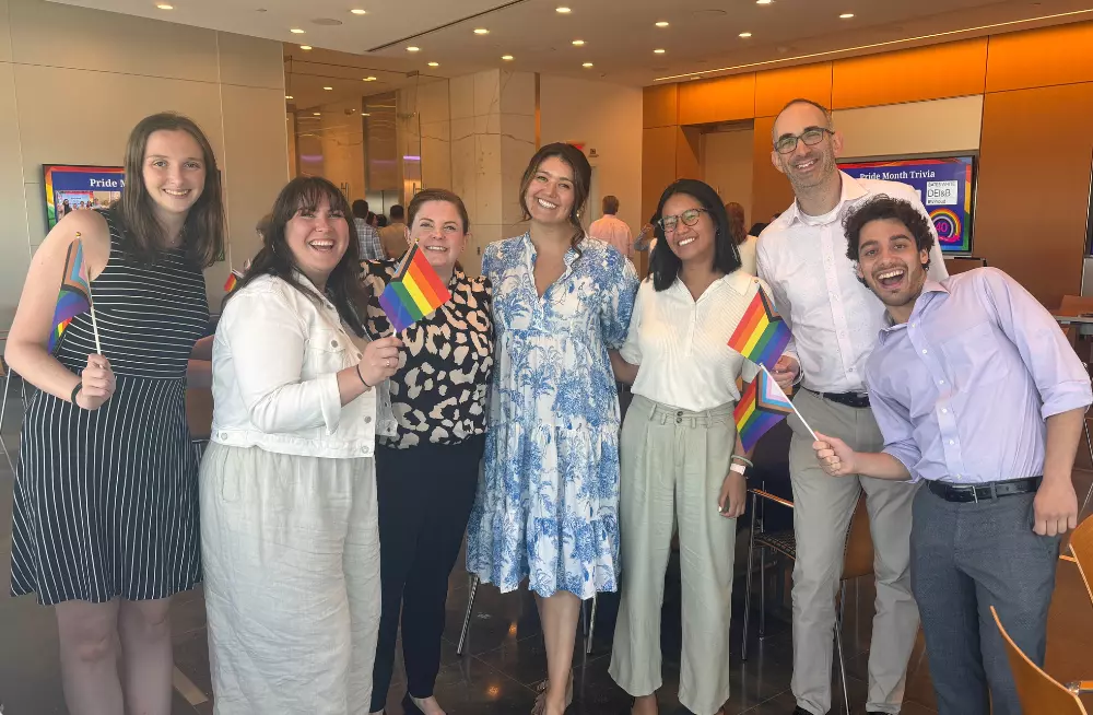 A group of 7 people (5 women, 2 men) stand together, smiling. Several of them are holding rainbow-colored Pride flags.