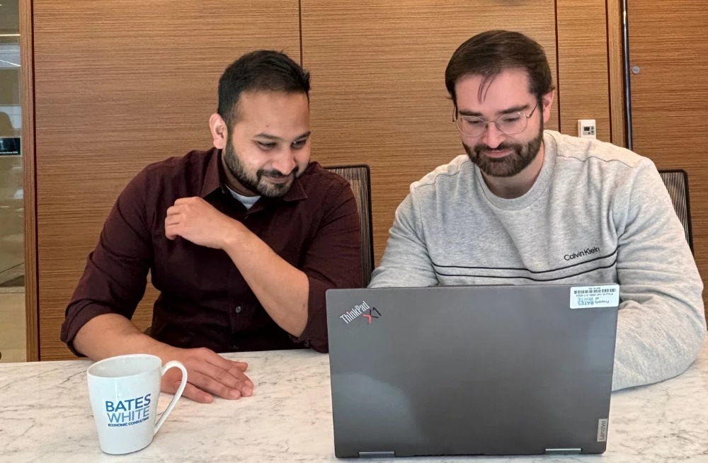 Two men sit behind a table, staring together at a laptop on said table. A coffee cup sits beside the laptop, with the Bates White logo.