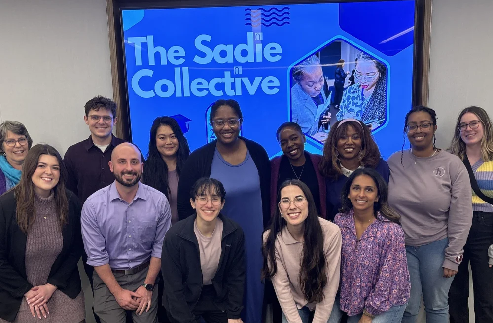 11 women and 2 men are grouped together, smiling at the camera. There are two rows of people, one in front of the other. A screen behind them is titled “The Sadie Collective.”