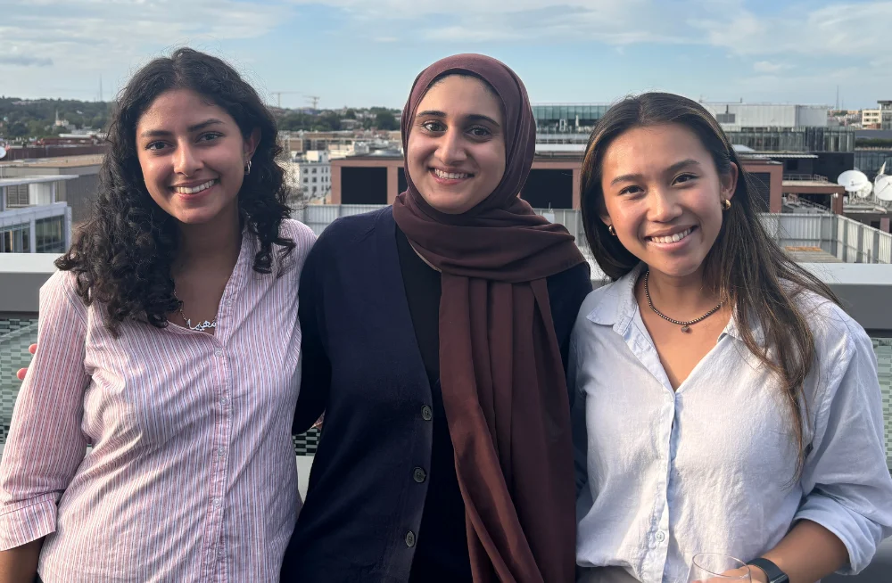 Three young women stand arm and arm on a rooftop. They are smiling at the camera. The background consists of an urban landscape and a partly cloudy sky.