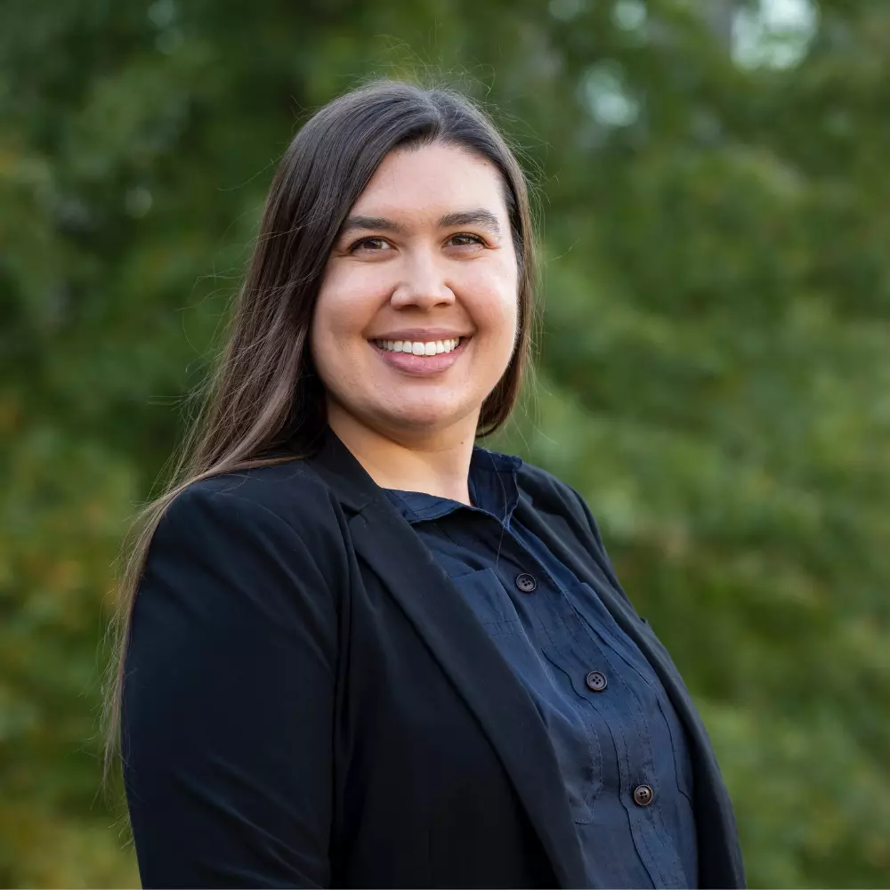A young woman stands outside, smiling at the camera, in front of a faded background of green trees.