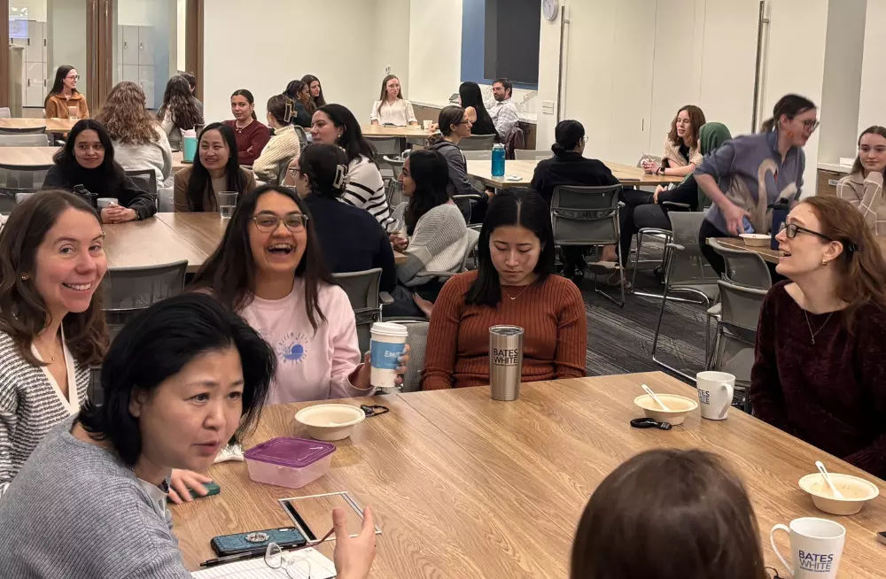 A group of women is gathered around a table. Some of in the middle of conversation, while others are smiling at the camera. Other tables behind them are also gathered around by people.
