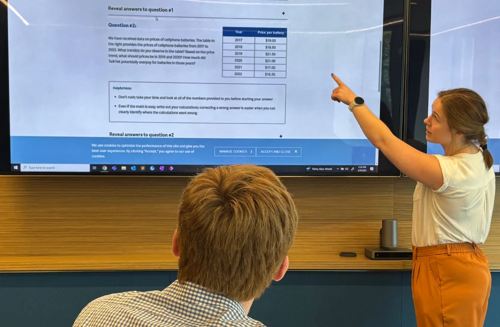 A young woman stands in front of a screen, pointing at a portion of it. The screen displays a portion of a case interview walkthrough on the Bates White website. A young man with his face away from the camera is seated, watching the screen.