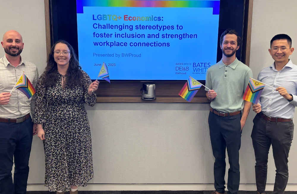 Three men and one woman stand to the sides of a screen, smiling at the camera. They are holding rainbow colored Pride flags. The screen is titled “LGBTQ+ Economics: Challenging stereotypes to foster inclusion and strengthen workplace connections.”
