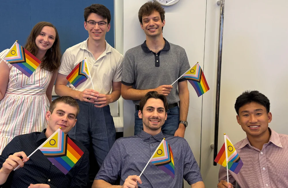 Two rows of people (3 people per row; one row is seated, one is standing) are smiling at the camera. They are holding rainbow colored Pride flags.