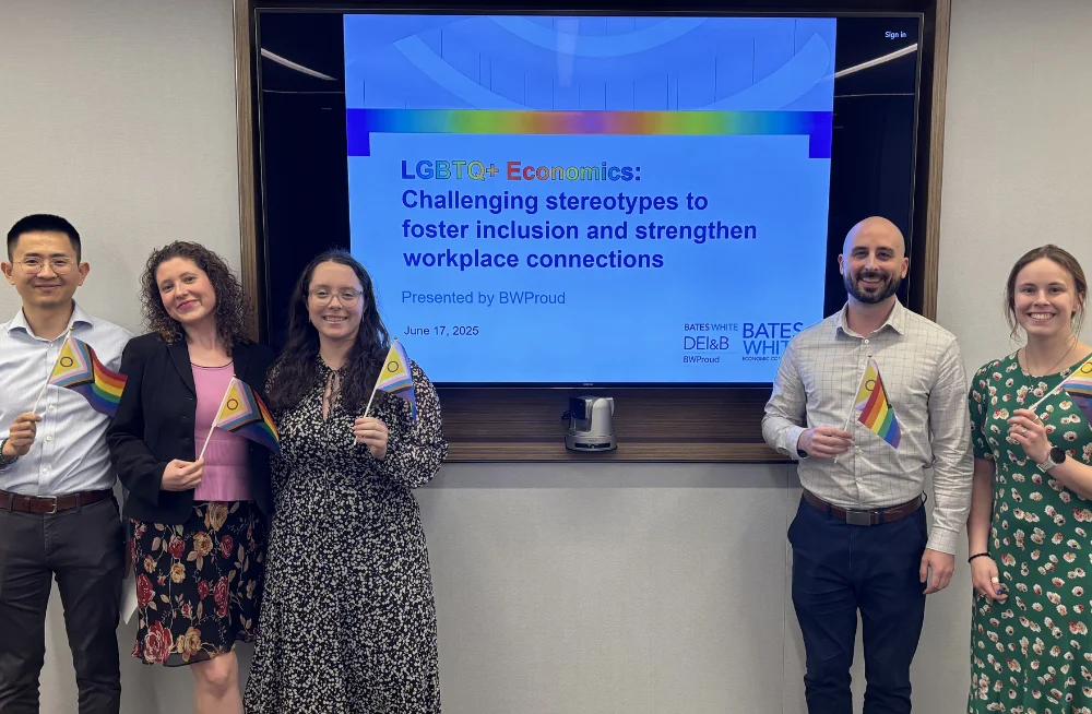 Two men and three women stand on either side of a large screen. They are smiling at the camera and are holding rainbow-colored Pride flags. The screen reads: “LGBTQ+ Economics: Challenging stereotypes to foster inclusion and strengthen workplace connectio