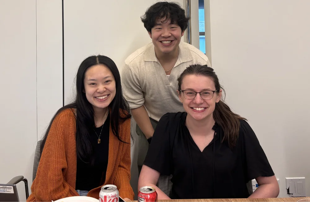 Two women sit behind a table, smiling at the camera. A man kneels behind and in between them, also smiling at the camera.