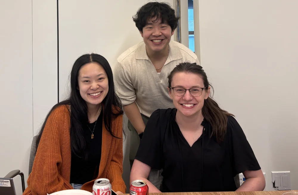 Two women are seated behind a table, smiling at the camera. In between and behind them is a standing man, who is also smiling.
