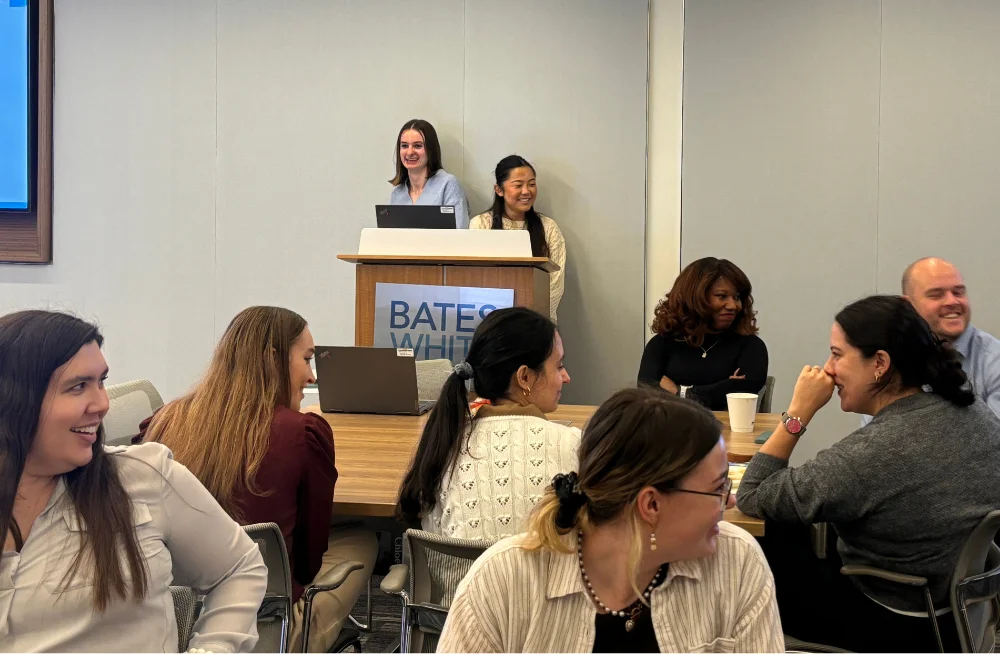 In the foreground, a group of smiling people (mostly young women; one young man) is seated around tables. In the background, two smiling women stand behind a speaking podium.