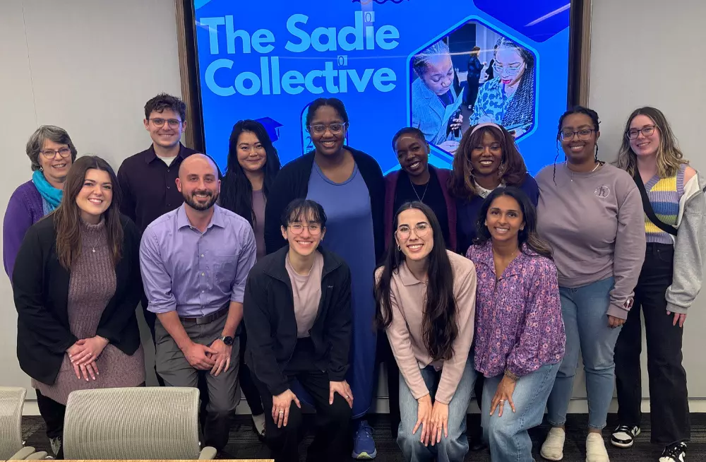 A group of mostly women with a few men pose together for a picture, smiling at the camera. Many of them are wearing purple clothing. A screen behind them is titled “The Sadie Collective.”