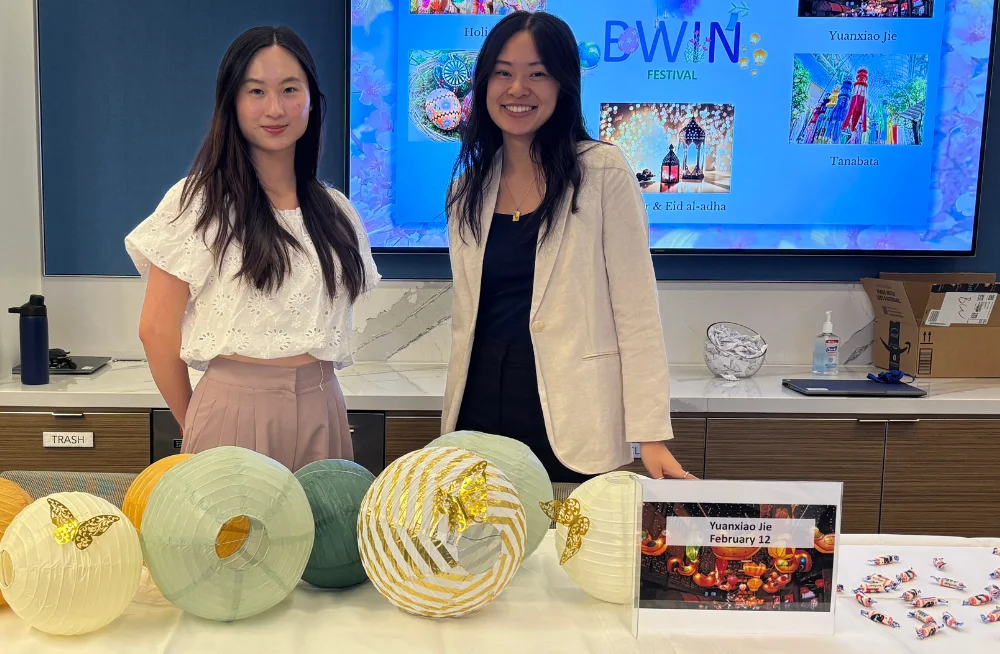 Two young women stand behind a table, smiling at the camera. The table has a sign reading “Yuanxiao Jie” on it. Items relating to the holiday, including wrapped candies and round paper ornaments, sit on the table. A screen behind them reads: “BWIN Festiva