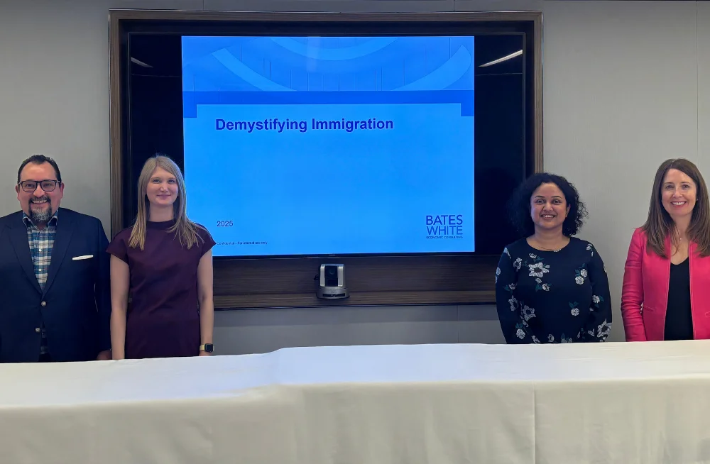 Three women and one man stand beside a large screen (two on the left and two on the right). They are smiling at the camera and are standing behind a table. The screen is titled “Demystifying Immigration.”