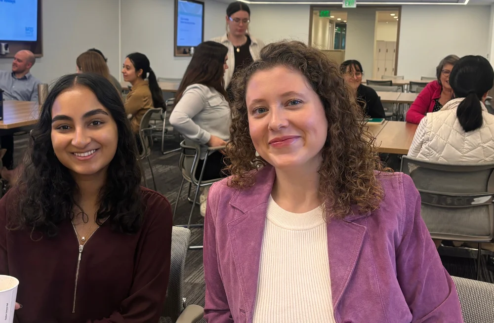 Two young women are seated next to each other, smiling at the camera. Behind them are two tables, each surrounded by a group of people in discussion.