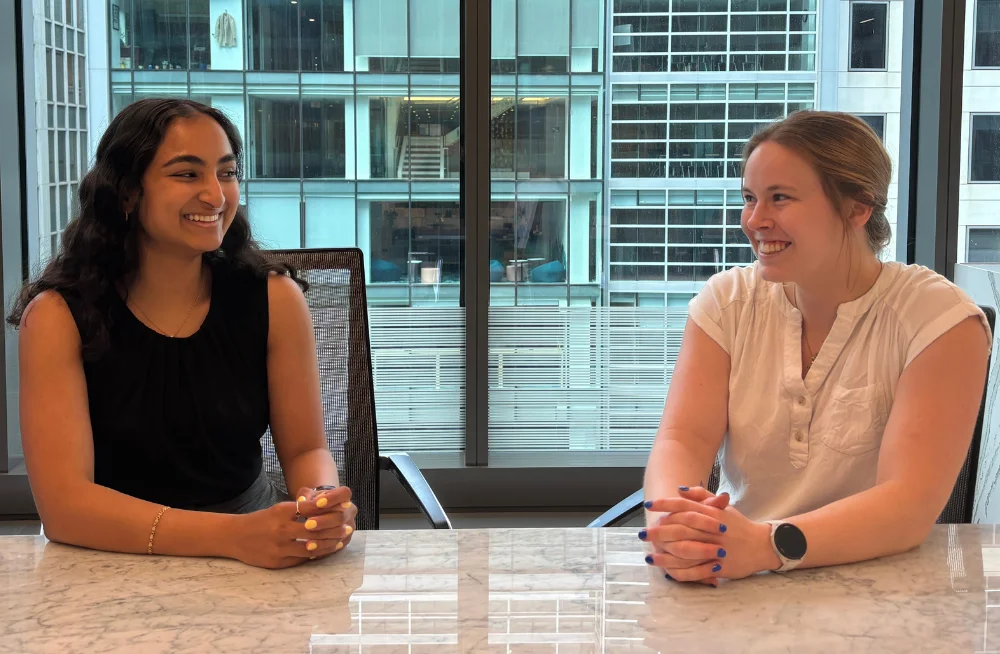 Two young women sit next to each other in front of a table. They are smiling at each other. A window behind them shows a tall office building.