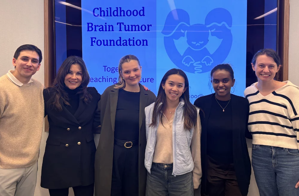 5 women and one man stand arm-in-arm together, smiling at the camera. A screen behind them is titled “Childhood Brain Tumor Foundation.”