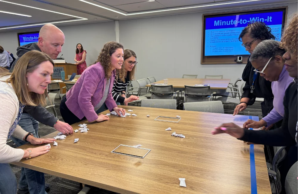Four adults lean on one side of a square table, facing 3 adults who are standing on the opposite side of the table. The group of four adults are trying to slide wrapped mints across the table. A screen behind them is titled “Minute-to-Win-It.”