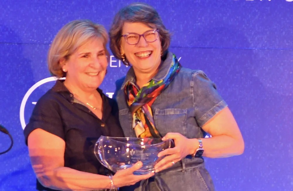 Two women stand in front of a blue backdrop, smiling at the camera. Together, they are holding a bowl-shaped award trophy.