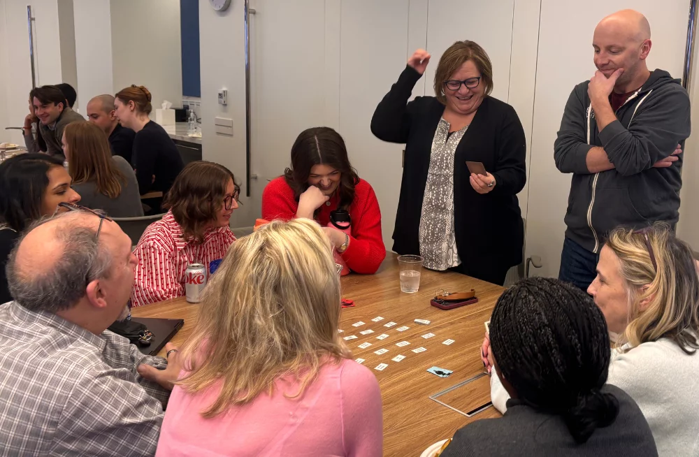 A group of men and women are gathered around a table—some standing and some sitting. One of the standing people smiles as she reads a card that she is holding. The table has around a dozen small paper slips on it.